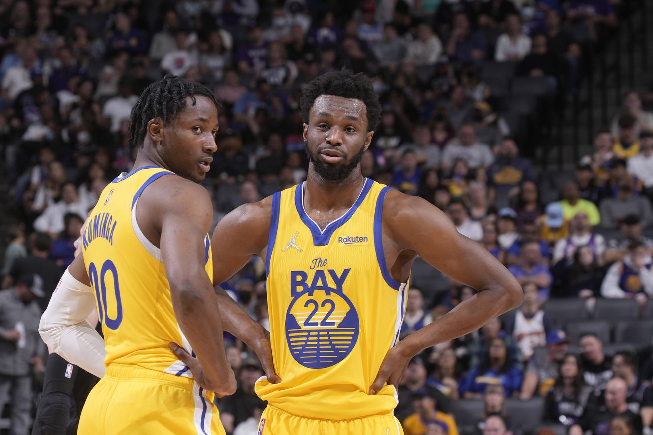 Andrew Wiggins with his hands on his hip, standing next to Jonathan Kuminga, looking over his shoulder.
