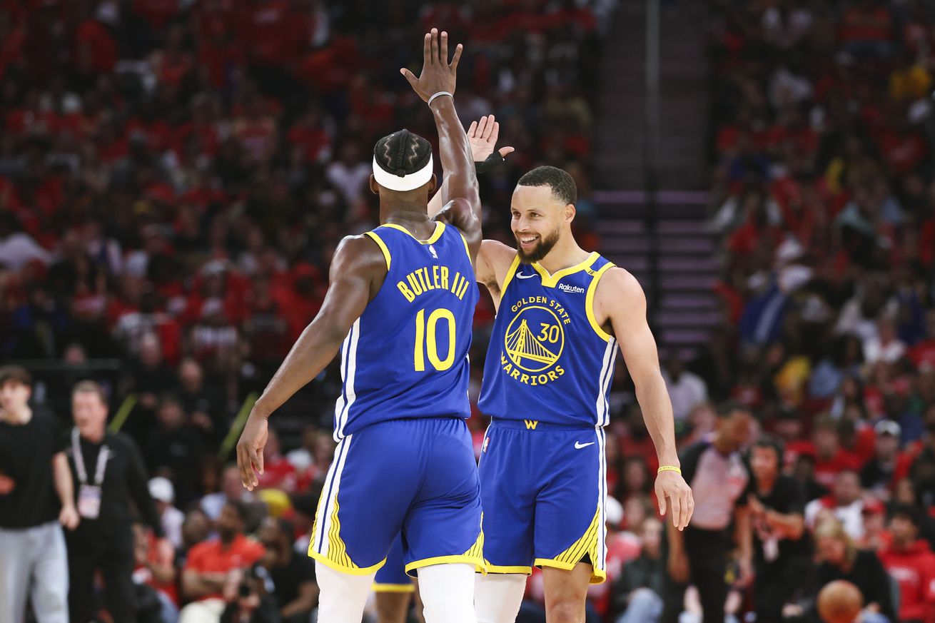 Steph Curry and Jimmy Butler III high-fiving on the court, with Curry facing the camera and Butler facing away from it.