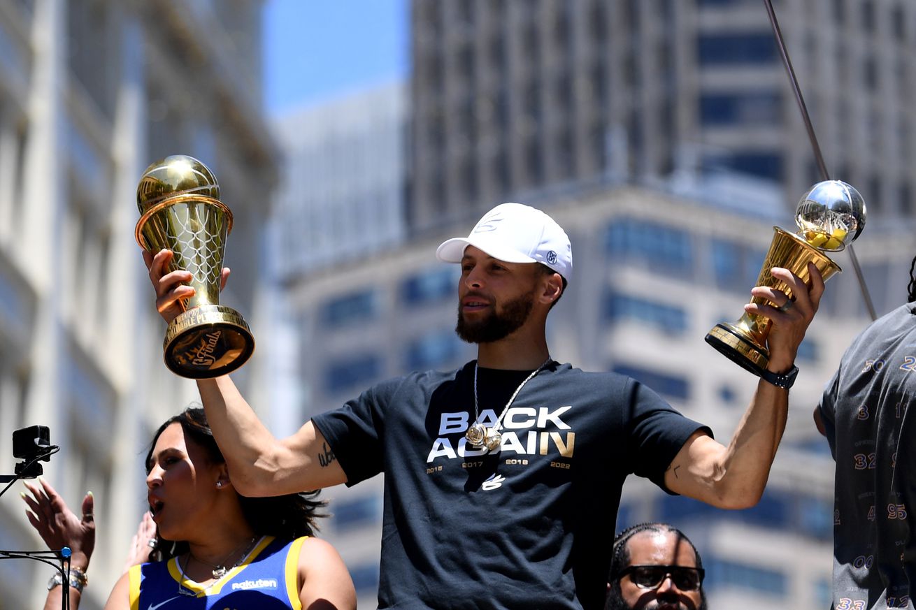 Steph Curry on a championship parade bus, holding the NBA Finals and NBA Finals MVP trophies. 