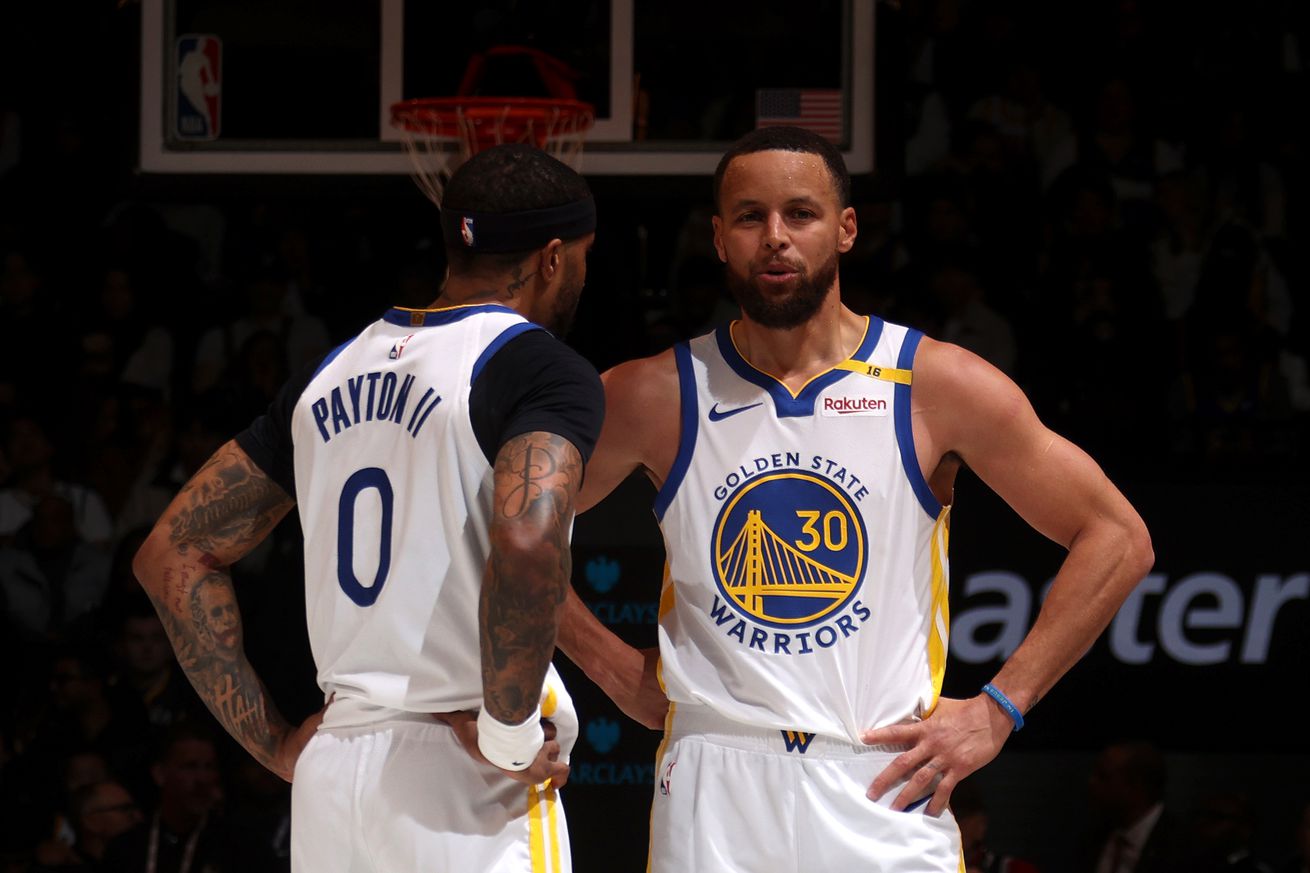 Steph Curry and Gary Payton II standing next to each other with hands on hips, talking.