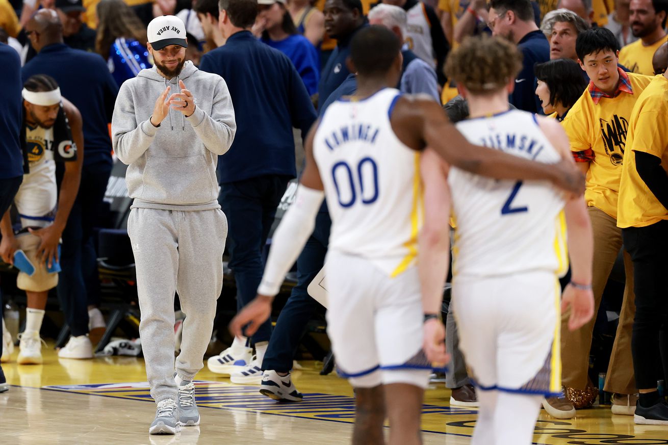 Steph Curry, in street clothes, clapping while Jonathan Kuminga and Brandin Podziemski walk off the court. 